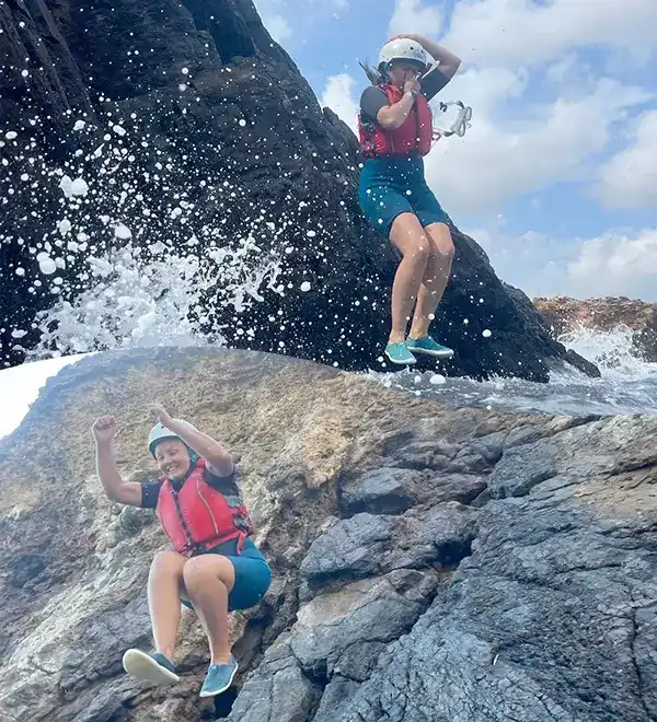 Mujer con casco y chaleco salvavidas saltando desde una roca hacia el mar con olas salpicando durante una sesion de coasteering en Ibiza.