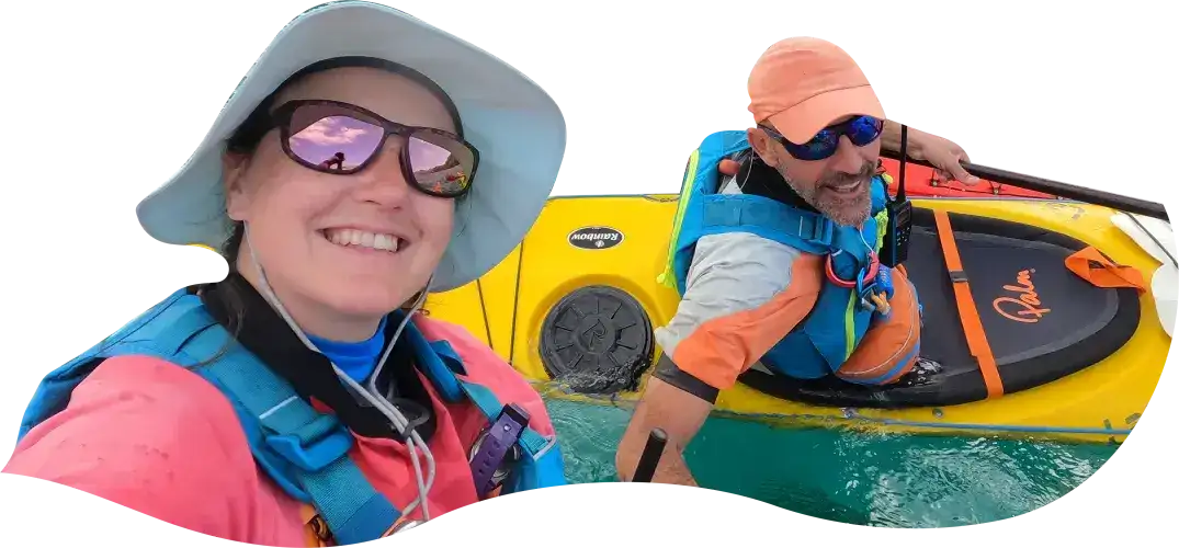 Dos guias expertos de kayak de mar en Ibiza sonriendo: mujer con sombrero, gafas de sol y equipamiento azul y rosa, y hombre con gorra naranja y chaleco salvavidas junto a un kayak amarillo.