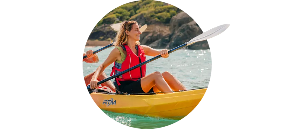 Mujer con chaleco salvavidas rojo remando en kayak amarillo por aguas tranquilas y turquesas con la costa verde de Ibiza de fondo durante la temporada de verano.