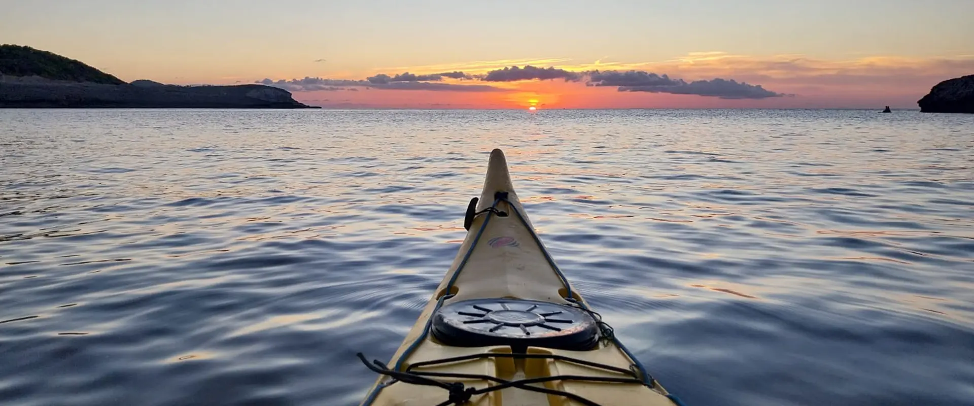 First-person view from a sea kayak bow gliding over calm waters toward a vibrant orange sunset with Ibiza's rocky coastline in the distance.